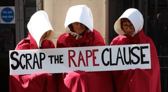 Three women dresses in Handmaid costumes holding a sign saying 'scrap the rape clause'